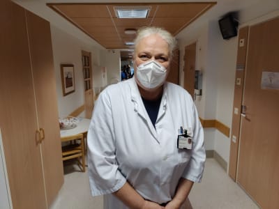 En kvinna med läkarrock står i en korridor. Hon har en ansiktsmask för munnen.