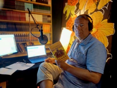 Ari Turunen äänikirjoja lukemassa, studiossa joka on sisutettu kirjoin ja kirjahyllyin.