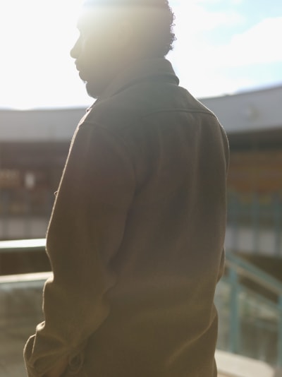 Ruskeisiin vaatteisiin pukeutunut parrakas mies seisoo ulkona. Miehestä näkyy siluetti, koska aurinko tulee hänen selkänsä takaa.
