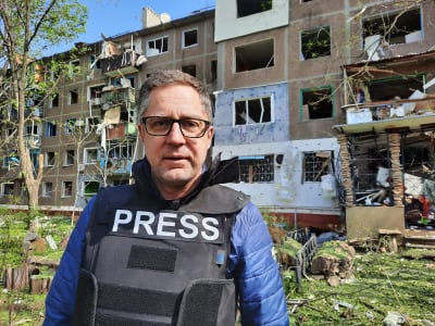 Antti Kuronen står framför ett bombskadat bostadshus med en säkerhetsväst på med texten "Press".