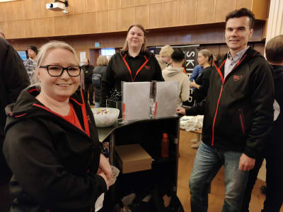 Tre personer står runt ett bord på en mässa. De heter Lotta Airo-Karlsson, Johanna Nyman och Henrik Tarvainen.