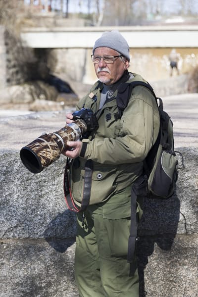 En äldre man med mörkgröna kläder håller i en kamera med ett stort objektiv.