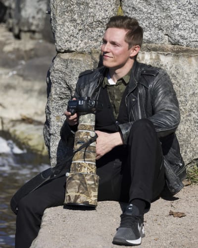 En ung, svartklädd man sitter på marken och lutar sig mot en stenvägg. I handen har han en kamera med ett stort objektiv.