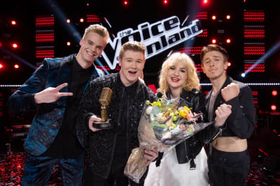 Tre unga män och en ung kvinna ler och tittar in i kameran efter finalen i The Voice of Finland. Vinnaren har en gyllene mikrofon och en blombukett i handen,