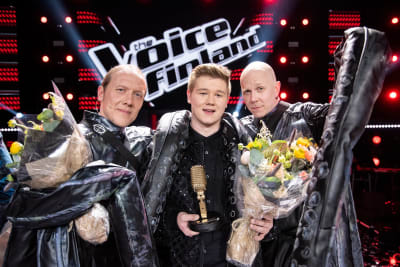 Tre män i svarta kläder håller i blombuketter och en pokal i form av en gyllene mikrofon. På väggen bakom dem står The Voice of Finland.