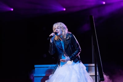 En ung kvinna med blont hår, stor vit klänning och svart läderjacka håller i en mikrofon och sjunger med ögonen slutna.