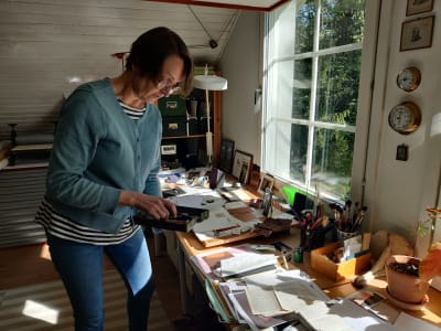 Maija Albrecht i sin konstnärsateljé. Hon håller i ett skrin med sina verktyg och står intill ett bord med arbetsredskap på.
