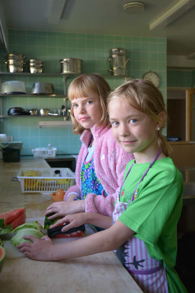 Två flickor med ljust hår skär vattenmelon och sallad. Den ena har på sig en ljusröd kohta och färggrant förkläde. Den andra har på sig en grön t-skjorta och färgrants förkläde. 