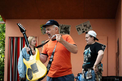 En man i orange t-skjorta och svart keps greppar en gitarr. I bakgrunden två ungdomar som tittar på.