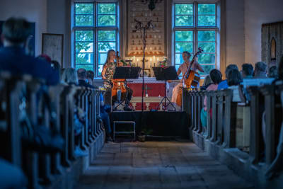 Två kvinnor i blommiga klänningar sitter längst fram i en gammal kyrka och spelar på  gamba och violone, två gamla stråkinstrument, i ljuset av stearinljus och det sista av dagens sol,