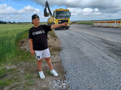 En pojke vid en landsväg pekar åt höger. En lastbild samt grävmaskin finns i bakgrunden.