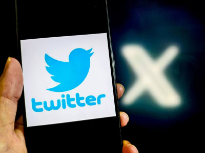 En hand håller upp en telefon med Twitters logo, i bakgrunden syns ett X.