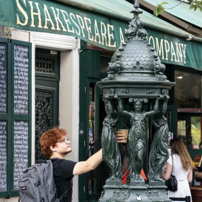 Shakespreare and company kirjakaupan edustalla reppuselkäinen nuori mies täyttää pahvimukia vanhasta veistoksin koristellusta vesipisteestä
