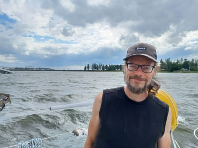 En skäggig man med keps på i sittbrunnen av en segelbåt. I bakgrunden syns Vasklot bro.