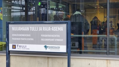 På en skylt utanför en byggnad står det Nuijamaa tull och gränsstation - persontrafikcentral på finska, svenska, engelska och ryska. I byggnaden bakom skylten kan man skymta människor genom ett stort fönster.