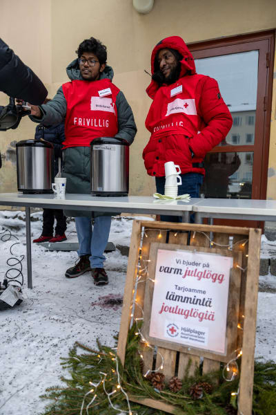 Två unga män iklädda vinterkläder och röd väst från Röda korset serverar varm dryck till hjälpbehövande som sträcker fram en hand. 