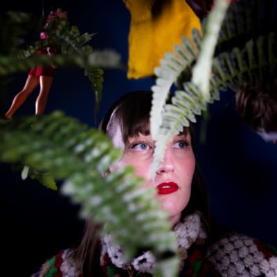 Elina Airio tummaa taustaa vasten, elinan kasvojen edessä pari saniaisen oksaa ja taustalla roikkuu nukkeja katosta.