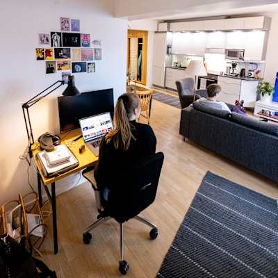 Insidan av en lägenhet där en person distansjobbar vid ett skrivbord och en annan ser på tv i bakgrunden.