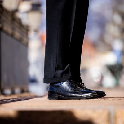 En anonymt klädd person i svarta kostym byxor står ute på en gata i svarta skor.