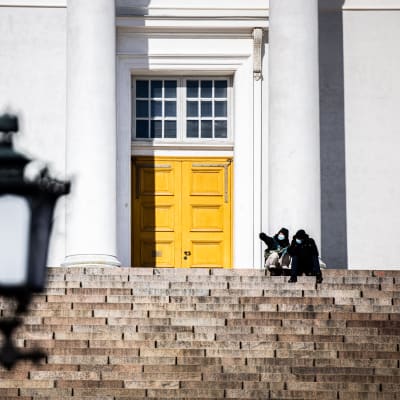 Två personer med munskydd sitter på Domkyrkan trappa i Helsingfors. 