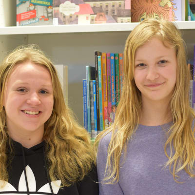 Två unga flickor framför en bokhylla med barnböcker