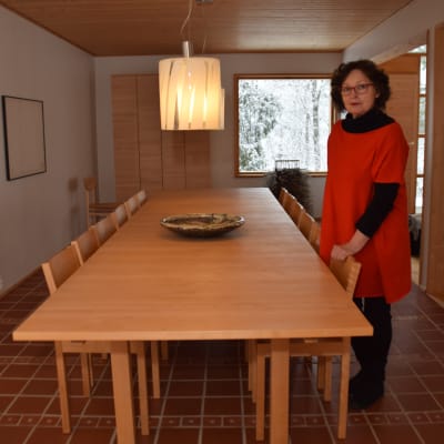En kvinna klädd i rött och svart och med röda glasögons tår vid ett långt köksbord. Hon heter Karin Widnäs.