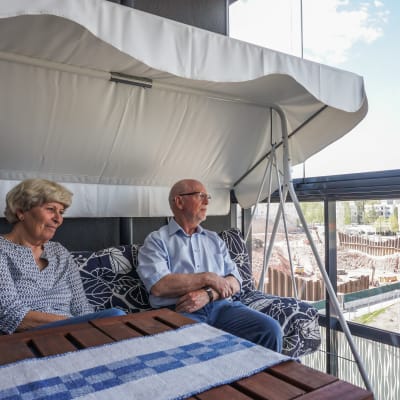 Britta (till vänster) och Matti Krank sitter på sin balkong, varifrån byggarbetsplatsen vid Lippulaiva syns.