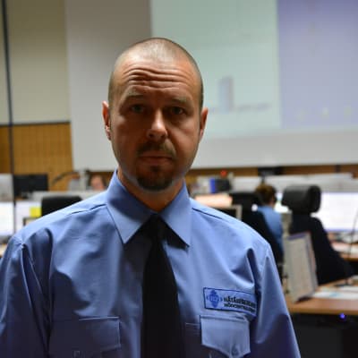 Matias Snellman bär en blå skjorta, svart slips. Han står med nödcentralens jourrum i bakgrunden. 