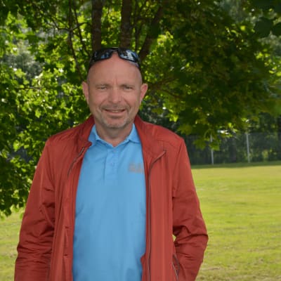 Dan Häggblom står i grönskan utomhus i blå skjorta och röd jacka och glasögon på huvudet.