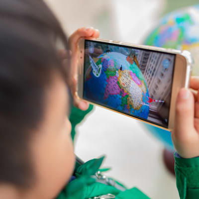 Kameran fokuserar på en mobilskärm som visar ett spel med en jordglob. I förgrunden ser vi ett suddigt barn som håller i mobilen.