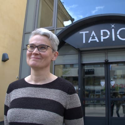 Raitapaitainen, harmaahiuksinen nainen seisoo Kino Tapio -elokuvateatterin pääoven edessä pihalla ja hymyilee.