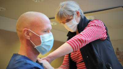 Kvinnlig sjukskötare ger spruta i axeln till en man i mörkblå sjukhusdräkt. Båda bär munskydd.