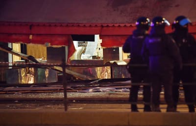 Kokokuvassa kolme kypäröin varustautunutta poliisia seisoo illalla kadulla. Taustalla kahvila, jonka ulkosivu on räjähtänyt.