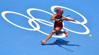 Naomi Osaka slår en boll med blått underlag och OS-ringarna i bakgrunden.