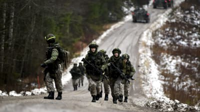 Beväringar deltar i en krigsövning i Kouvola. De bär på vapen och går upp för en backe på en landsväg med skog på båda sidorna.