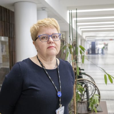 Kvinna klädd i mörkblå blus ser in i kameran. I bakgrunden sjukhusmiljö.