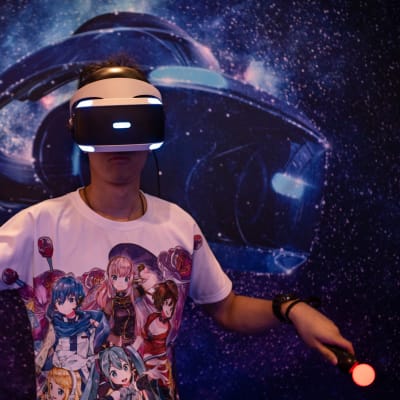 En man med VR-glas och kontroller i händerna ses spela ett spel. Han har på sig en t-shirt med anime-figurer.