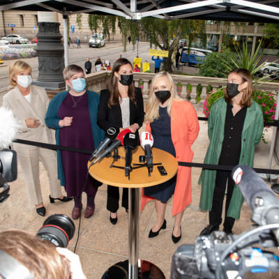 Partiledarna i Sanna Marins regering står bakom ett runt bord fyllt med mikrofoner och fotograferas.