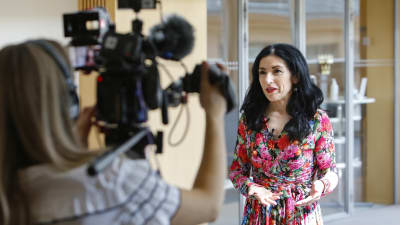 Amineh Kakabaveh talar framför en kamera. Hon är klädd i en blommig klänning och har rött läppstift på sig.