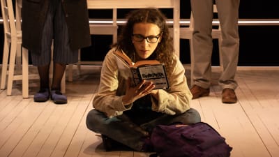En ung kvinna sitter på golvet och läser en bok. Bakom henne står hennes föräldrar.