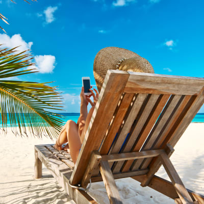 En kvinna i en solstol på en strand sitter och petar på en smarttelefon.