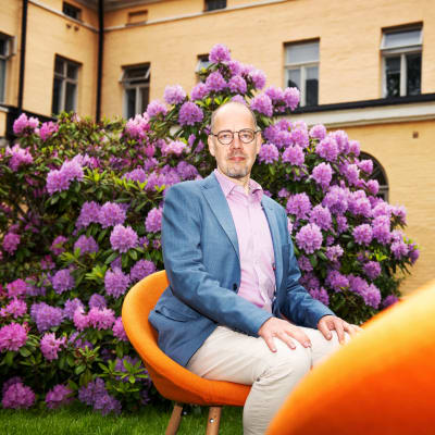 Mies istuu kukkapuskan edessä ulkona oranssilla tuolilla.