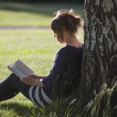 Nainen istuu nurmikolla, lukee kirjaa ja nojaa koivuun