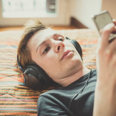 En kvinna med kort hår ligger på en säng och lyssnar på musik. Hon har hörlurar på sig och tittar på sin smarttelefon. 