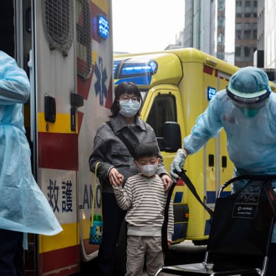 Sjukvårdare på gatan i blåa plastdräkter framför en ambulans. Bredvid står en kvinna och ett barn med ansiktsmasker.