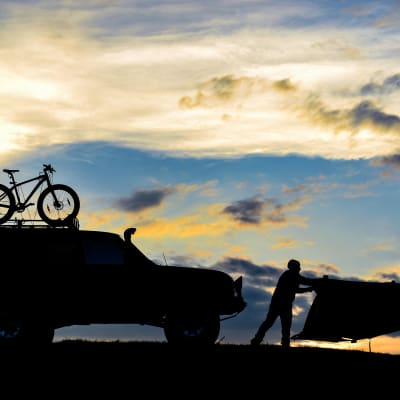 Pilvinen taivas ja silhuettina näkyvä jeeppi, polkupyörä ja teltta
