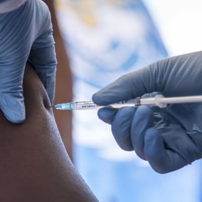 En hand med en blå gummihandske håller i en vaccinspruta som tryckts in i en persons arm.