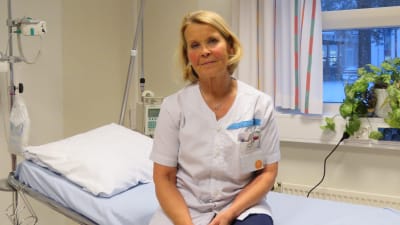 En kvinna i sköterskekläder sitter på en brits i ett rum på ett sjukhus.