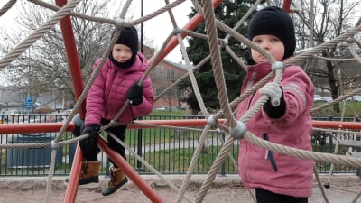 Astrid och Ebba Tuominen, två flickor i rosa jackor och svarta mössor, leker i en klätterställning med metallvajrar.