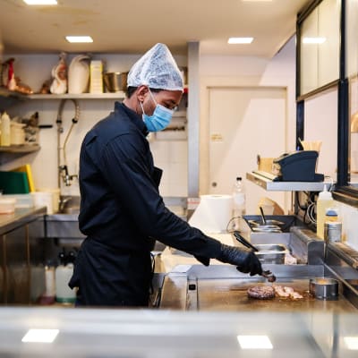En man i munskydd står i ett restaurangkök och tillreder kött.